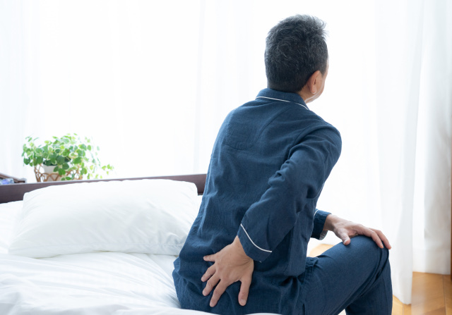 パジャマ姿でベッドの端に座り腰に手を当てる男性