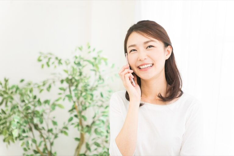 白いトップスで観葉植物の前に立ちスマートフォンで通話する女性