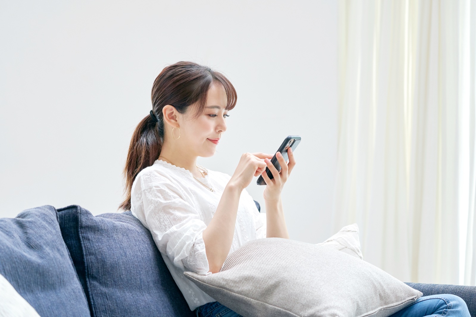 ソファにもたれて背中を丸め、スマートフォンを見る女性