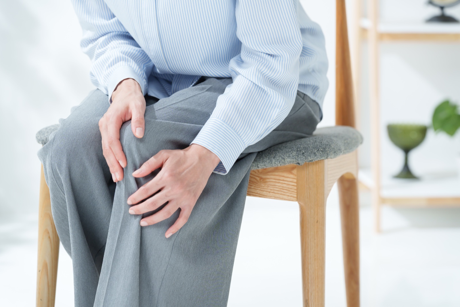 木の椅子に浅く腰かけ両手で片膝を押さえるグレーのパンツの人