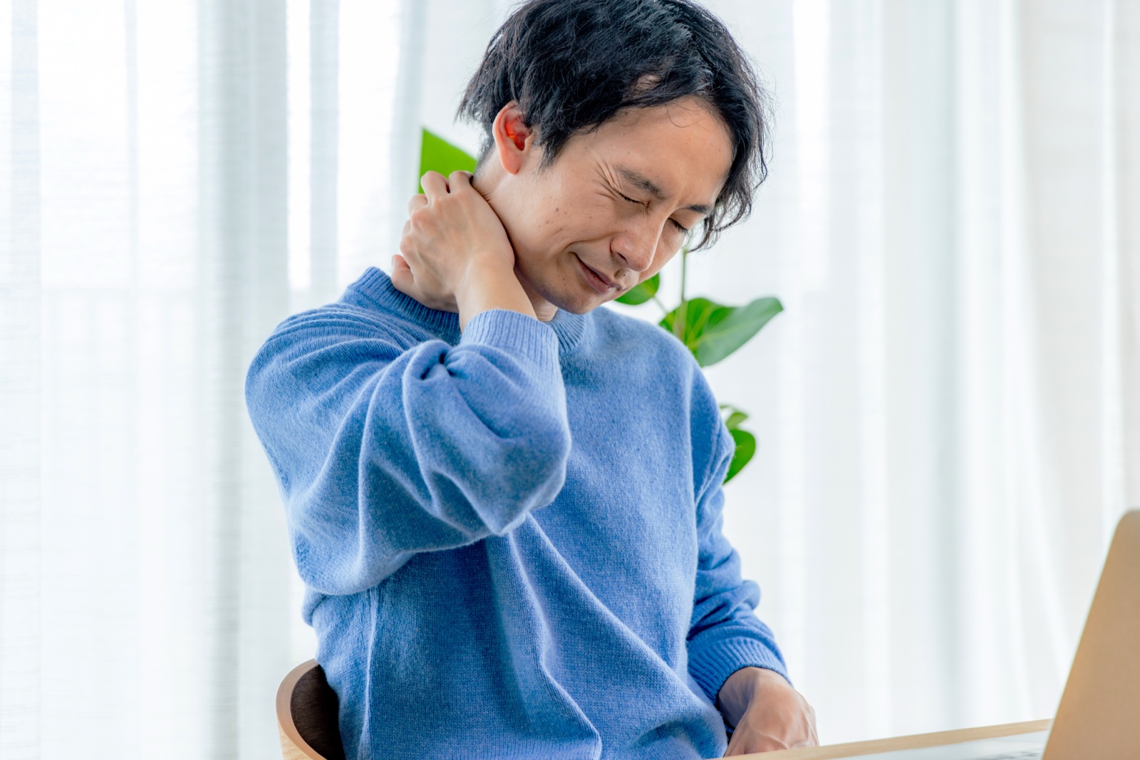デスクで首の後ろに手を当てて顔をしかめる青いセーターの男性
