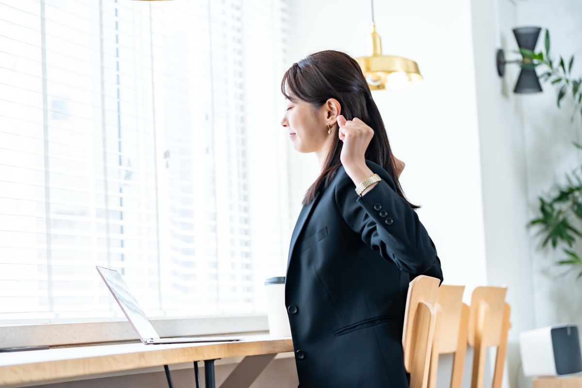 窓辺のデスクでひじを後ろに引いて肩を動かすスーツの女性