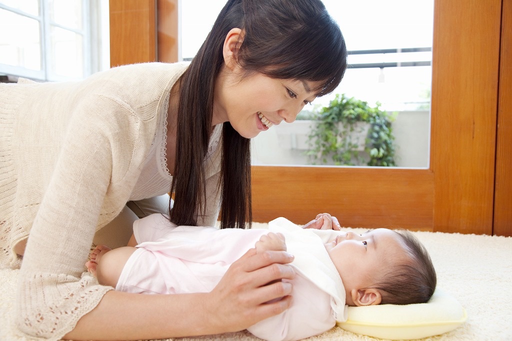 床に寝た赤ちゃんに前かがみで顔を近づけてあやす女性